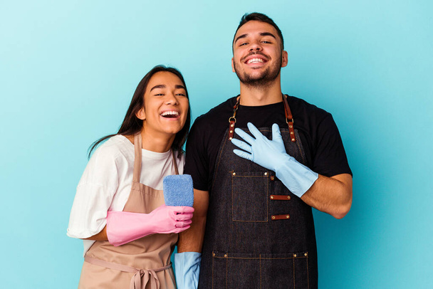 joven mixta raza pareja limpieza casa aislado en azul fondo risas fuera ruidosamente mantener la mano en pecho. - Foto, Imagen