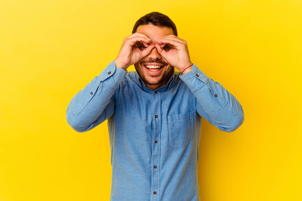 Young caucasian man isolated on yellow background showing okay sign over eyes - Fotoğraf, Görsel