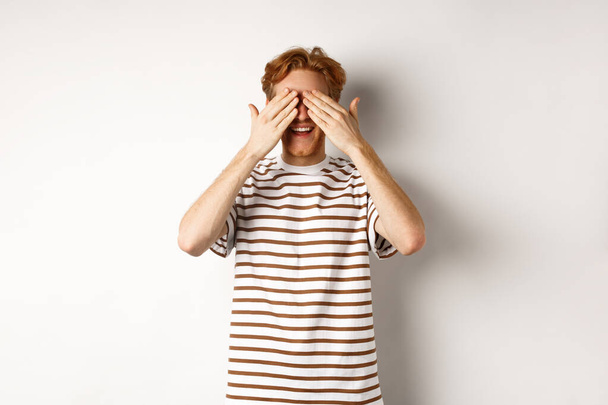 Smiling man cover eyes and waiting for surprise, standing in t-shirt against white background - Photo, Image