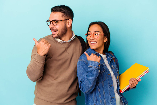Young mixed race student couple isolated on blue background points with thumb finger away, laughing and carefree. - Photo, image