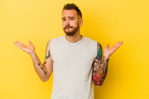 Young tattooed caucasian man isolated on yellow background confused and doubtful shrugging shoulders to hold a copy space. - Photo, Image
