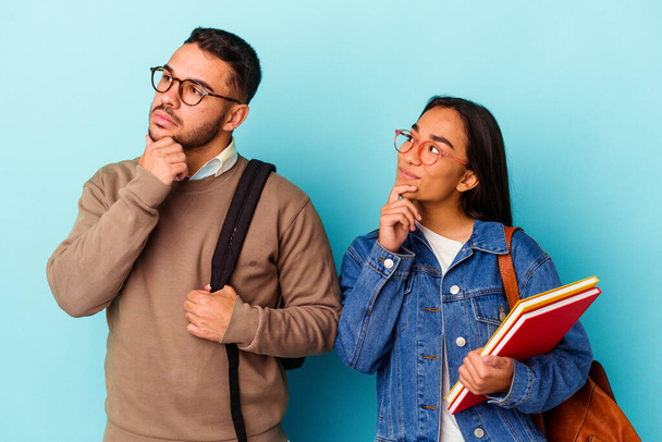 Jeune couple mixte étudiant isolé sur fond bleu regardant latéralement avec une expression douteuse et sceptique. - Photo, image