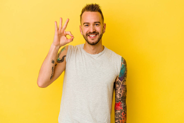 Jeune homme caucasien tatoué isolé sur fond jaune joyeux et confiant montrant ok geste. - Photo, image