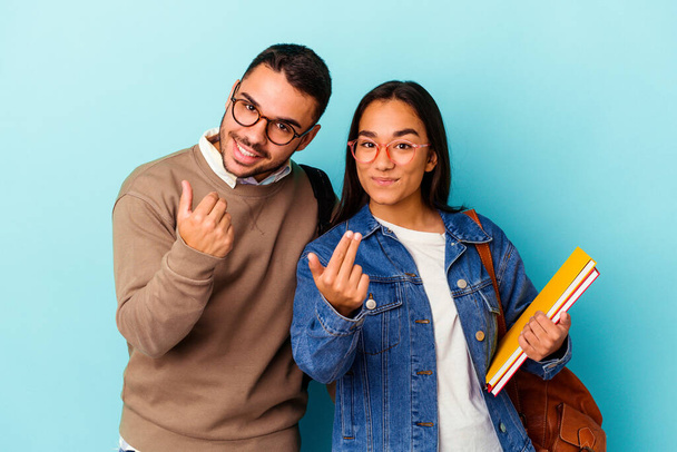 Mladý smíšený závod studentský pár izolované na modrém pozadí ukazuje prstem na vás, jako by pozvání přijít blíž. - Fotografie, Obrázek