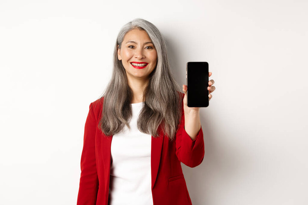 Iäkäs liikenainen punaisessa bleiserissä, näyttää tyhjän älypuhelimen näytön ja hymyilee, osoittaa mobiilisovellus, valkoinen tausta - Valokuva, kuva