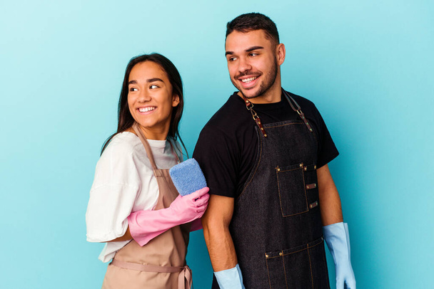 Young mixed race couple cleaning home isolated on blue background looks aside smiling, cheerful and pleasant. - Photo, Image