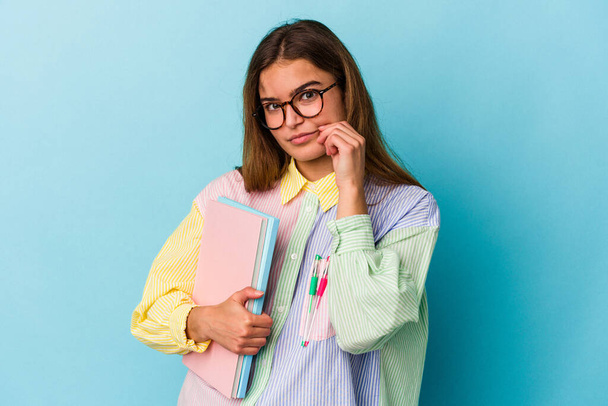 Νεαρή καυκάσια φοιτήτρια που κρατάει βιβλία απομονωμένα σε μπλε φόντο με δάχτυλα στα χείλη κρατώντας ένα μυστικό. - Φωτογραφία, εικόνα