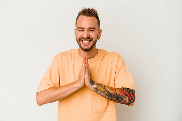 Young tattooed caucasian man isolated on white background holding hands in pray near mouth, feels confident. - Foto, immagini