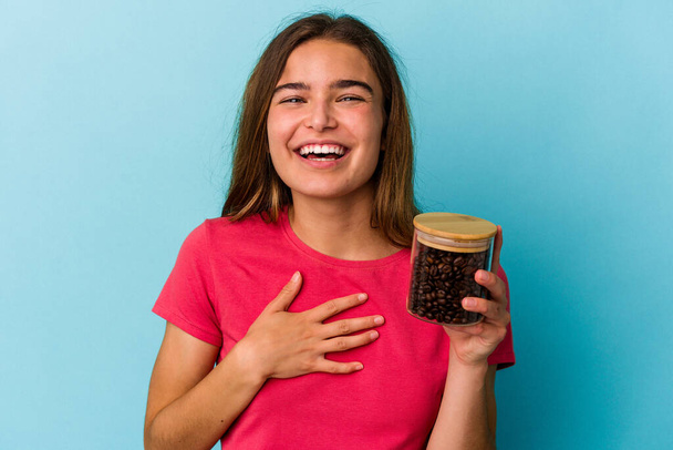 Νεαρή λευκή γυναίκα που κρατάει ένα βάζο καφέ απομονωμένο σε μπλε φόντο γελάει δυνατά κρατώντας το χέρι στο στήθος. - Φωτογραφία, εικόνα