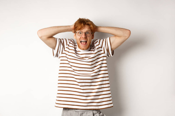 Frustrated redhead man panicking, screaming and holding hands on head, standing on white background - Photo, Image