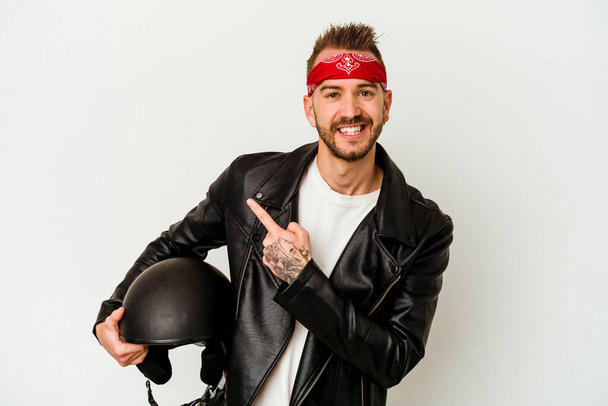 Giovane motociclista tatuato uomo caucasico in possesso di un casco isolato su sfondo bianco sorridente e indicando da parte, mostrando qualcosa a spazio vuoto. - Foto, immagini