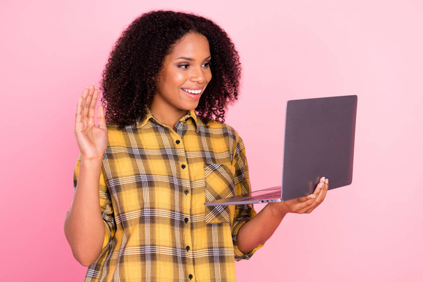 Foto de adorable linda mujer de piel oscura vestida camisa a cuadros hablando dispositivo moderno agitando brazo aislado de color rosa fondo - Foto, imagen