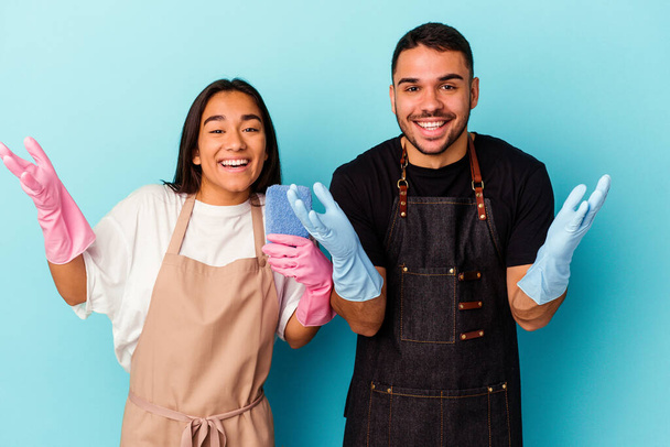 Junges gemischtes Paar, das isoliert auf blauem Hintergrund zu Hause putzt, erhält eine angenehme Überraschung, ist aufgeregt und hebt die Hände. - Foto, Bild