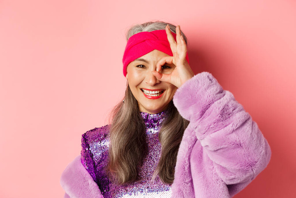 Moda. Primo piano della donna anziana asiatica alla moda sorridente, mostrando OK segno sopra gli occhi e guardando felice la fotocamera, in piedi su sfondo rosa - Foto, immagini