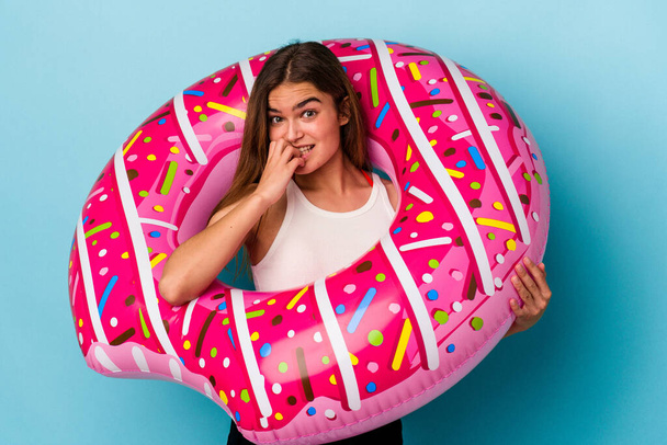 Young caucasian woman with inflatable donut isolated on blue background biting fingernails, nervous and very anxious. - Photo, Image