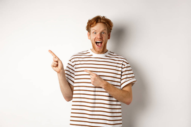 Cheerful man with red hair checking out promo offfer, pointing left at logo and smiling at camera, standing over white background - Photo, Image