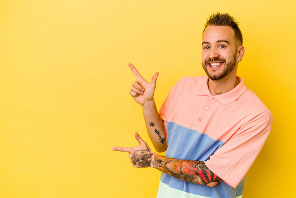 Jovem tatuado caucasiano homem isolado no fundo amarelo animado apontando com os dedos indicador de distância. - Foto, Imagem