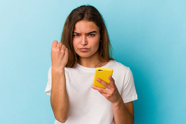 Young caucasian woman holding a mobile phone isolated on blue background showing fist to camera, aggressive facial expression. - Photo, Image