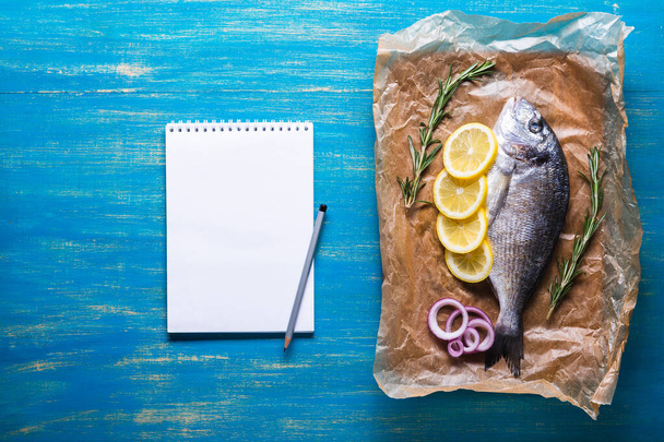Frischer roher Dorado-Fisch auf Kochpapier mit Gewürzen und einem Notizbuch für Rezept oder Speisekarte. Draufsicht auf blauem Hintergrund. - Foto, Bild