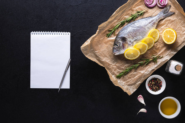 Roh Dorado auf Kochpapier mit Gewürzen und einem Notizbuch für Rezept oder Speisekarte. Gesundes Essen, Meeresfrüchte. Draufsicht auf schwarzem Hintergrund. - Foto, Bild