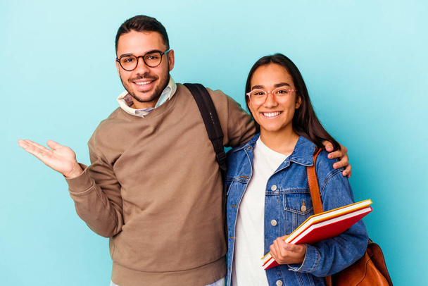 Joven pareja de estudiantes de raza mixta aislada sobre fondo azul mostrando un espacio de copia en una palma y sosteniendo otra mano en la cintura. - Foto, Imagen