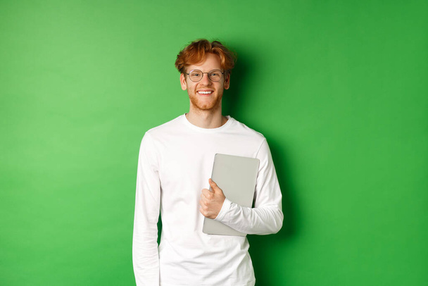 Handsome young man with red hair, wearing glasses and long-sleeve t-shirt, holding laptop on green background - Foto, afbeelding