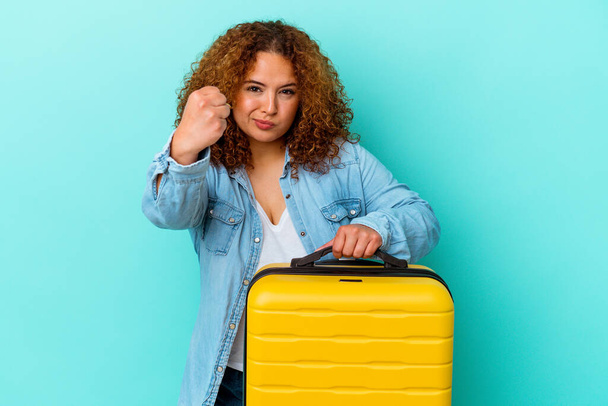 Giovane viaggiatore latino donna curvy in possesso di una valigia isolata su sfondo blu mostrando pugno alla macchina fotografica, espressione facciale aggressiva. - Foto, immagini