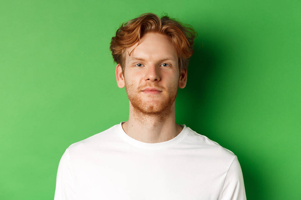 Nahaufnahme eines jungen Mannes mit rotem, unordentlichem Haar und Bart, der in die Kamera schaut, vor grünem Hintergrund stehend - Foto, Bild