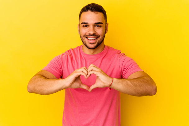 Молодий кавказький чоловік ізольований на жовтому тлі посміхаючись і демонструючи форму серця руками
. - Фото, зображення