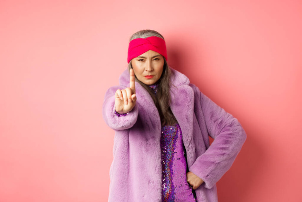 Koncepcja mody i zakupów. Poważna azjatycka seniorka pokazująca nie tak szybki gest, potrząsająca wyciągniętym palcem, aby zatrzymać lub ostrzec, patrząca zdeterminowana w kamerę, ubrana w stylowe fioletowe ubrania - Zdjęcie, obraz