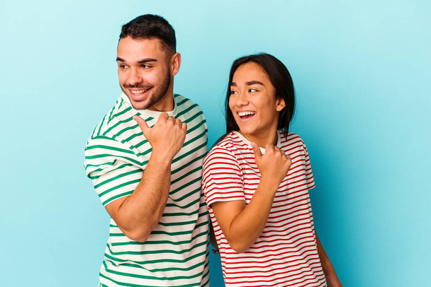Young mixed race couple isolated on blue background points with thumb finger away, laughing and carefree. - Photo, Image