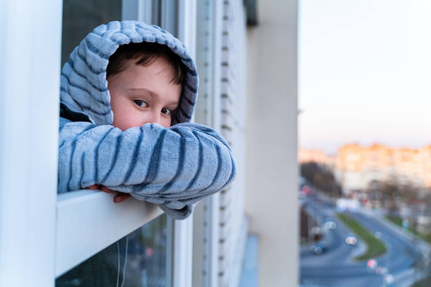 Quarantine. Little boy stands and look out of a window bored. Longing for fresh air and walks along the street. Forced home during quarantine due to the coronavirus pandemic - Foto, Imagen