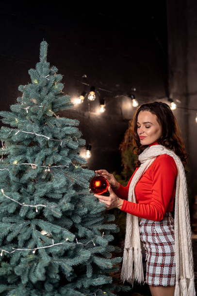 Pretty woman posing to the camera while looking at the ball dreamy standing near the fir tree. Red sweater on a nice girl. New year decorations. Holidays concept - Fotografie, Obrázek