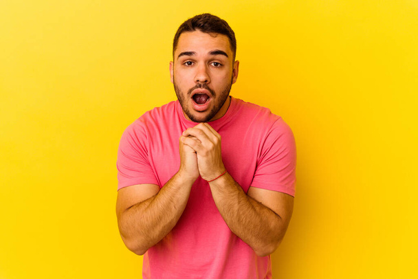 Young caucasian man isolated on yellow background praying for luck, amazed and opening mouth looking to front. - Photo, Image
