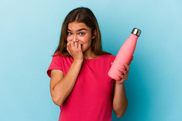Молодая белая женщина, держащая бутылку воды на голубом фоне, кусает ногти, нервничает и очень беспокоится. - Фото, изображение