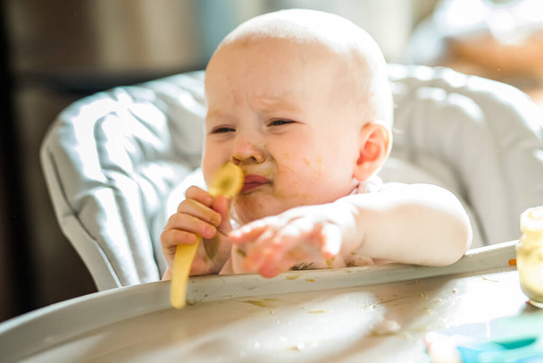 6 maanden oud meisje thuis In de kinderstoel lekker eten thuis. Grappige baby met bevuilde gezonde broccoli puree gezicht houdt lepel in de handen - Foto, afbeelding