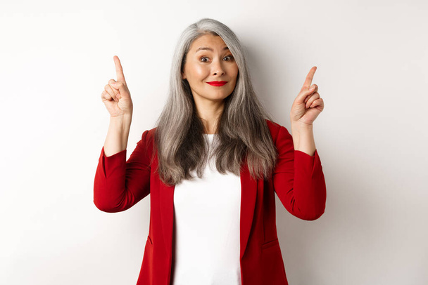 Elegant asian senior lady in red blazer and makeup, pointing fingers up and smiling, showing advertisement, white background - Fotografie, Obrázek