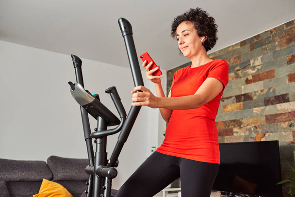 Femme faisant de l'exercice à domicile sur vélo elliptique à l'aide d'un téléphone portable - Photo, image