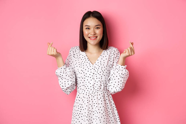 Schöne asiatische Frau in Kleid mit koreanischem Herz Zeichen und lächelnd, steht auf romantischem rosa Hintergrund - Foto, Bild