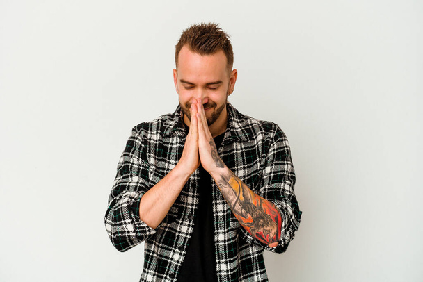 Young tattooed caucasian man isolated on white background holding hands in pray near mouth, feels confident. - Foto, Bild