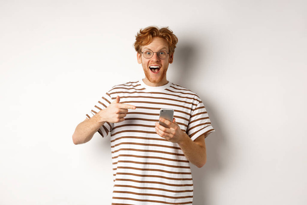 技術と電子商取引の概念.携帯電話でオンラインプロモーションをチェックアウト素晴らしい赤毛の男,スマートフォンや笑顔で指を指して,白い背景 - 写真・画像