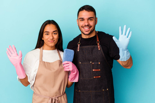 Молодая смешанная раса пара уборки дома изолированы на синем фоне улыбаясь весело показывая номер пять с пальцами. - Фото, изображение