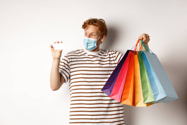 Pandemic and special offer concept. Happy redhead man in face mask showing shopping bags and plastic credit card, standing over white background - Fotoğraf, Görsel