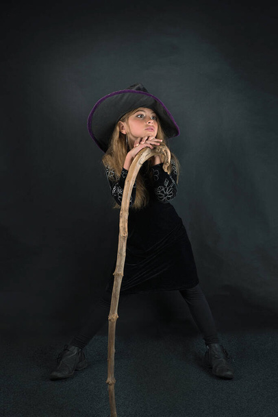 Een meisje op een donkere achtergrond in een heksenkostuum en in een brede hoed met een staf voor Halloween. - Foto, afbeelding