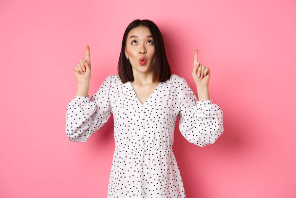 Cute korean girl in beautiful dress saying wow, looking and pointing fingers up, intrigued in promo offer, standing over pink background - Photo, Image
