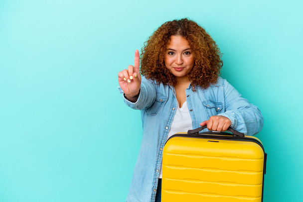Young latin traveler curvy woman holding a suitcase isolated on blue background showing number one with finger. - Photo, Image