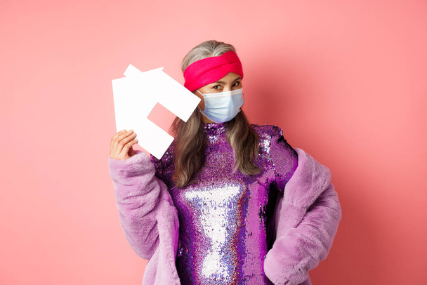 Covid-19 a realitní koncept. Cool a stylové asijské starší žena v diskotéce šaty a lékařská maska ukazující papírenský dům, stojící nad růžové pozadí - Fotografie, Obrázek