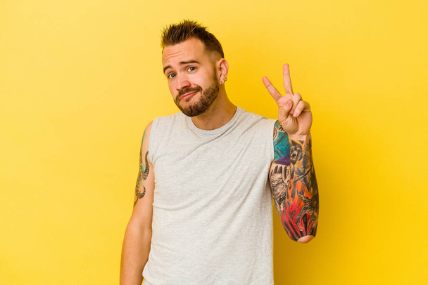 Jeune homme caucasien tatoué isolé sur fond jaune joyeux et insouciant montrant un symbole de paix avec les doigts. - Photo, image