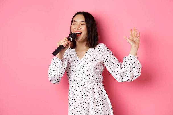 Szczęśliwa azjatycka kobieta tańcząca i śpiewająca w mikrofonie, występująca na karaoke, stojąca nad różowym tle - Zdjęcie, obraz