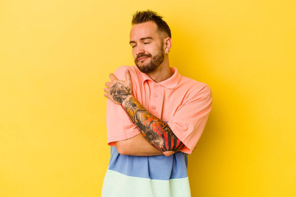 Joven hombre caucásico tatuado aislado en abrazos de fondo amarillo, sonriente despreocupado y feliz. - Foto, Imagen